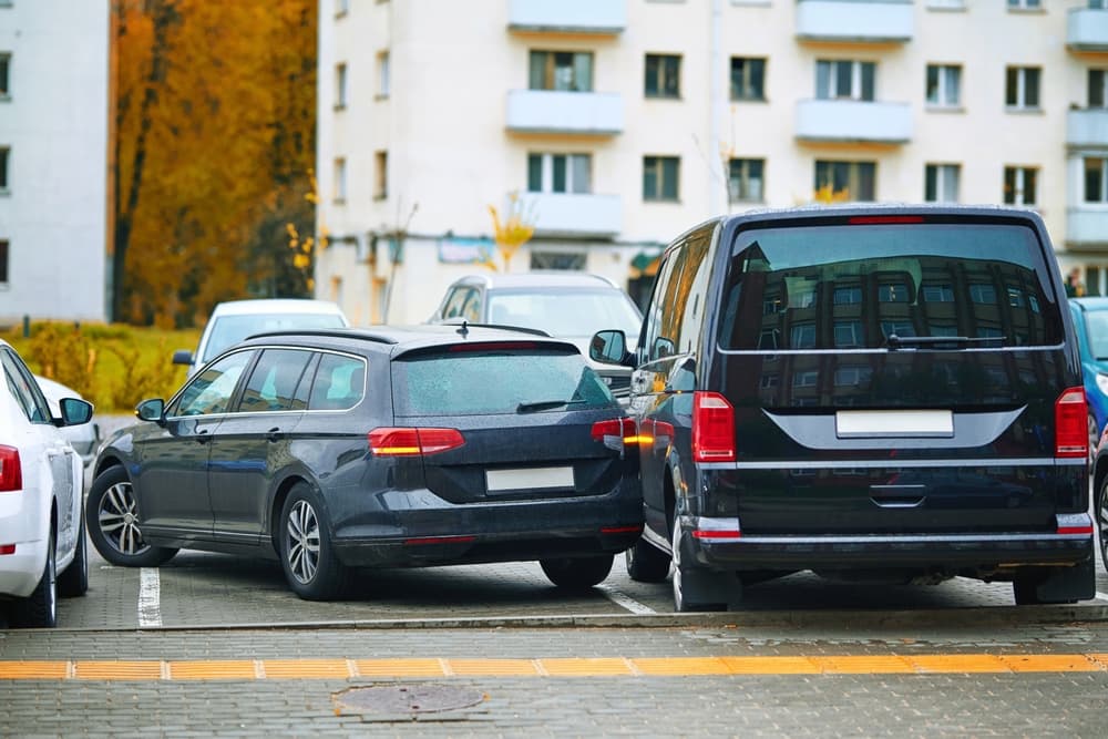 A black sedan reverses into a parked van in a cramped parking lot, causing a collision. The accident results from limited visibility and distracted driving.