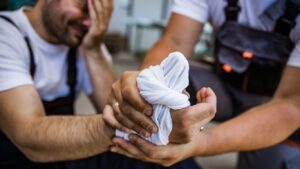 A blue-collar worker wraps a bandage around his injured coworker’s hand, illustrating a burn injury claim in Alabama.