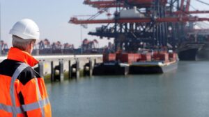 A dockworker in safety gear on a shipping pier with cargo containers in the background. Dockworker injury claims fall under the Longshore and Harbor Workers Act.