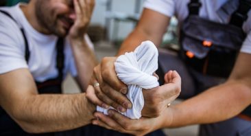 A blue-collar worker wraps a bandage around his injured coworker’s hand, illustrating a burn injury claim in Alabama.