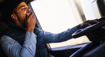 A tired truck driver rubbing his eyes behind the wheel of a semi, illustrating truck driver hours of service fatigue rules.